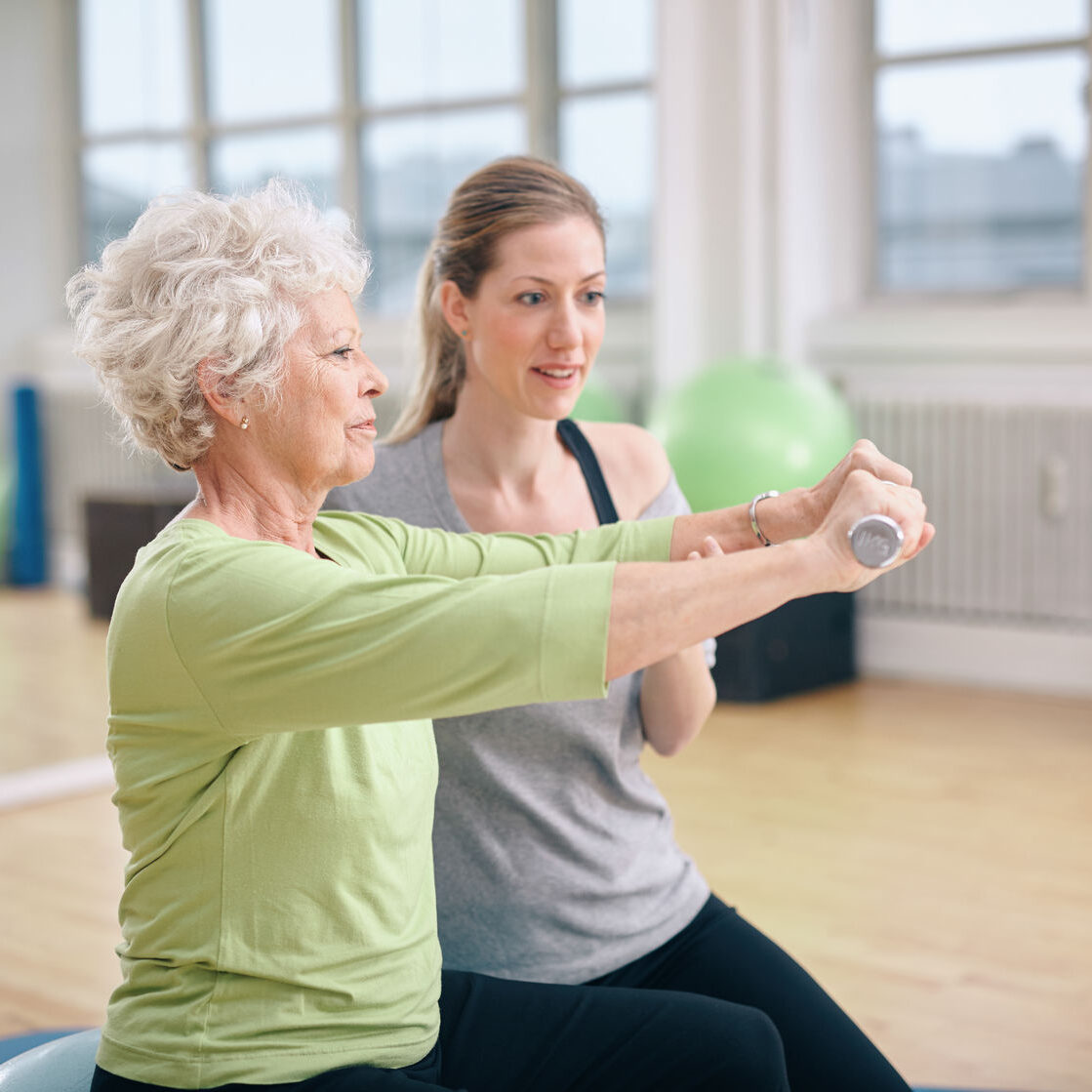 34387158 - senior woman being assisted by instructor in lifting dumbbells at gym. senior woman training in the gym with a personal trainer at rehab.